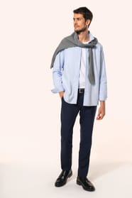 Jeune homme aux cheveux châtains courts portant une chemise bleu clair en popeline à manches longues, ouverte sur un T-shirt blanc et rehaussée d’un pull gris clair noué sur les épaules. Confectionnée en coton peigné double retors 80x2, elle se distingue 