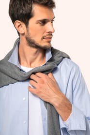 Jeune homme aux cheveux châtains courts portant une chemise bleu clair en popeline à manches longues, ouverte sur un T-shirt blanc et rehaussée d’un pull gris clair noué sur les épaules. Confectionnée en coton peigné double retors 80x2, elle se distingue 