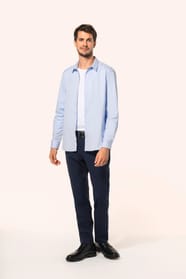 Jeune homme aux cheveux châtains courts portant une chemise bleu clair en popeline à manches longues, ouverte sur un T-shirt blanc. Confectionnée en coton peigné double retors 80x2, elle se distingue par sa coupe ajustée avec pinces dans le dos, ses finit