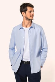 Jeune homme aux cheveux châtains courts portant une chemise bleu clair en popeline à manches longues, ouverte sur un T-shirt blanc. Confectionnée en coton peigné double retors 80x2, elle se distingue par sa coupe ajustée avec pinces dans le dos, ses finit