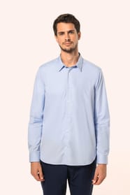 Jeune homme aux cheveux châtains courts portant une chemise bleu clair en popeline à manches longues, ouverte sur un T-shirt blanc. Confectionnée en coton peigné double retors 80x2, elle se distingue par sa coupe ajustée avec pinces dans le dos, ses finit