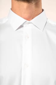 Jeune homme aux cheveux châtains courts portant une chemise blanche en twill à manches longues, confectionnée en coton double retors 80x2 de qualité supérieure. Sa coupe ajustée, structurée par deux pinces dans le dos, se distingue par des finitions raffi
