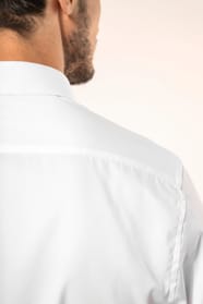 Jeune homme aux cheveux châtains courts portant une chemise blanche en twill à manches longues, confectionnée en coton double retors 80x2 de qualité supérieure. Sa coupe ajustée, structurée par deux pinces dans le dos, se distingue par des finitions raffi
