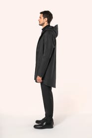 Jeune homme aux cheveux châtains courts portant une veste de pluie unisexe noire en tissu imperméable 5000 mm, dotée d’une capuche à visière et de coutures thermosoudées. Confectionnée avec une enduction polyuréthane à l’extérieur et une doublure polyeste