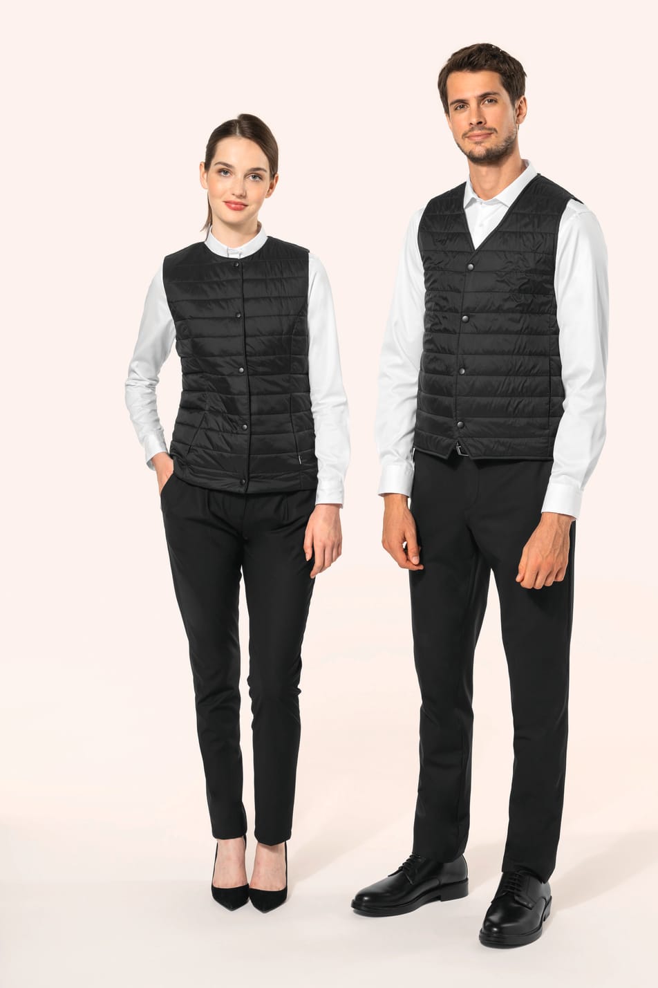 Jeune femme aux cheveux châtains longs attachés en queue de cheval et jeune homme aux cheveux châtains courts portent un bodywarmer léger noir, pensé pour se glisser aisément sous une veste. Confectionnés en polyamide avec un rembourrage intérieur en Thin