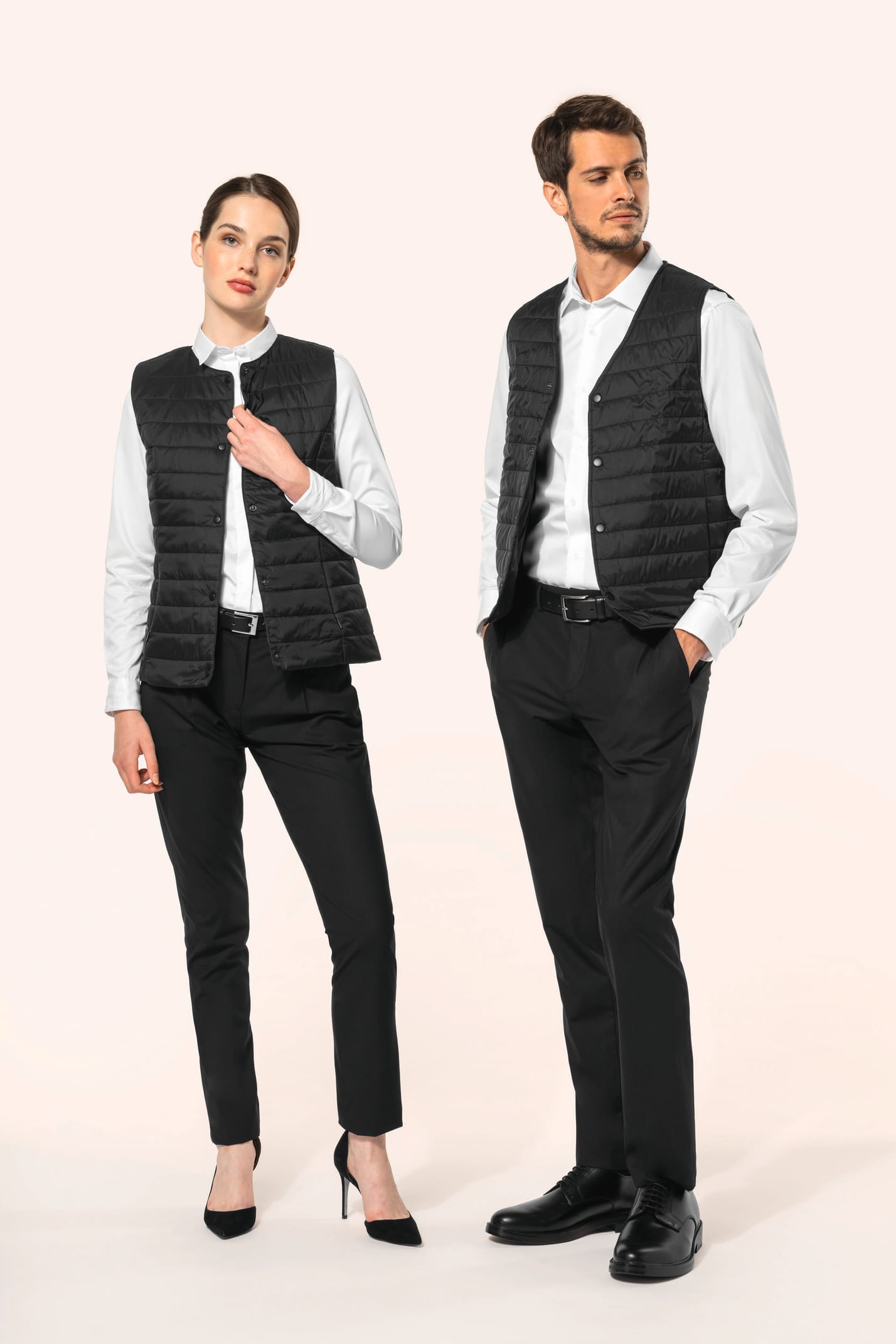 Jeune femme aux cheveux châtains longs attachés en queue de cheval et jeune homme aux cheveux châtains courts portent un bodywarmer léger noir, pensé pour se glisser aisément sous une veste. Confectionnés en polyamide avec un rembourrage intérieur en Thin