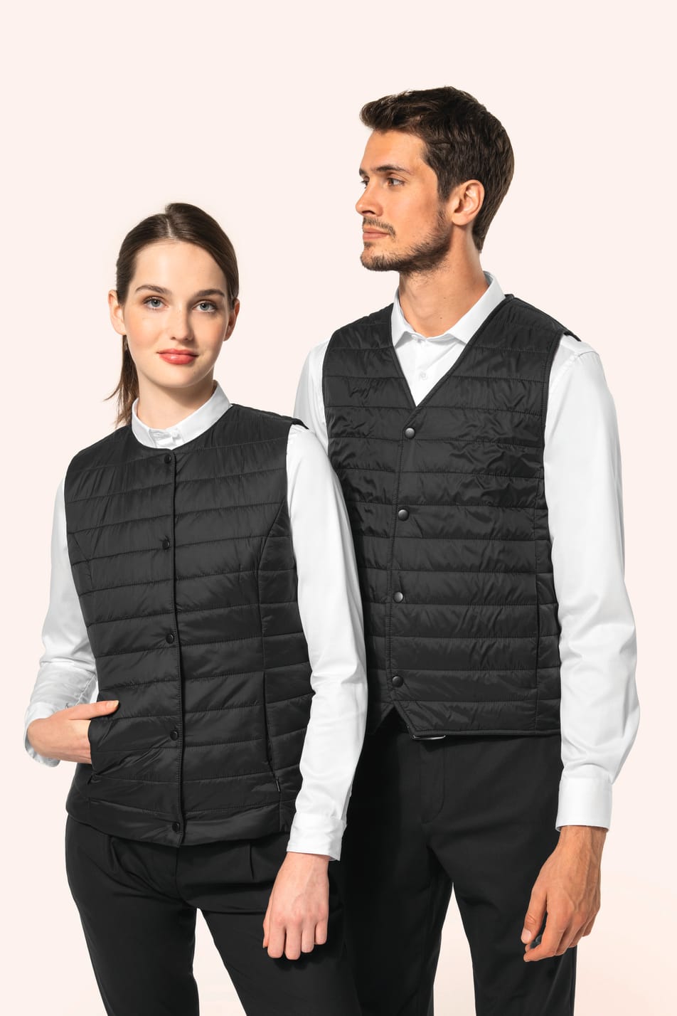 Jeune femme aux cheveux châtains longs attachés en queue de cheval et jeune homme aux cheveux châtains courts portent un bodywarmer léger noir, pensé pour se glisser aisément sous une veste. Confectionnés en polyamide avec un rembourrage intérieur en Thin