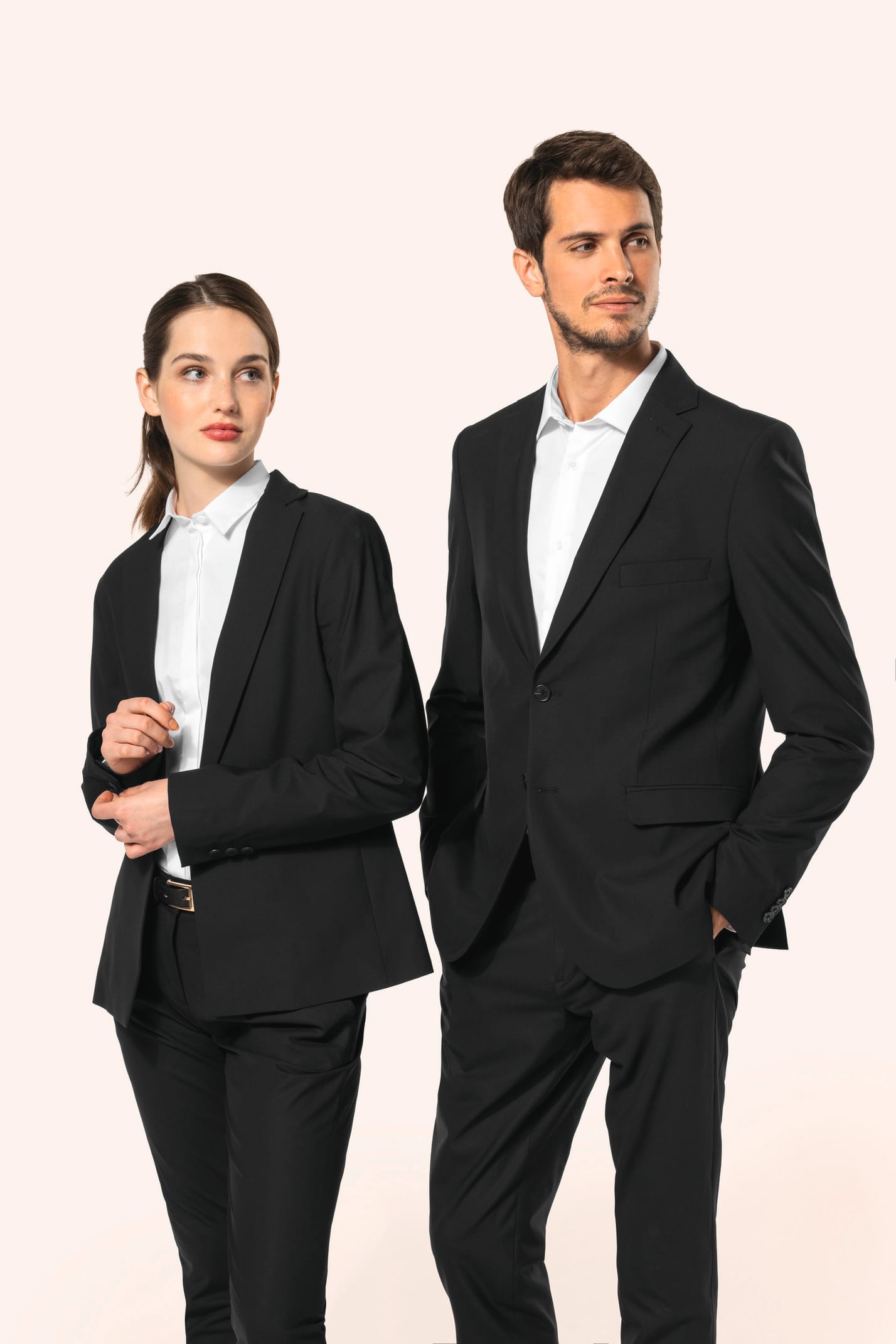 Jeune homme aux cheveux châtains courts et jeune femme aux cheveux châtains longs attachés en queue de cheval composent un duo élégant en noir et blanc. Ils portent chacun une veste noire à la coupe ajustée, associée à une chemise blanche, un pantalon noi