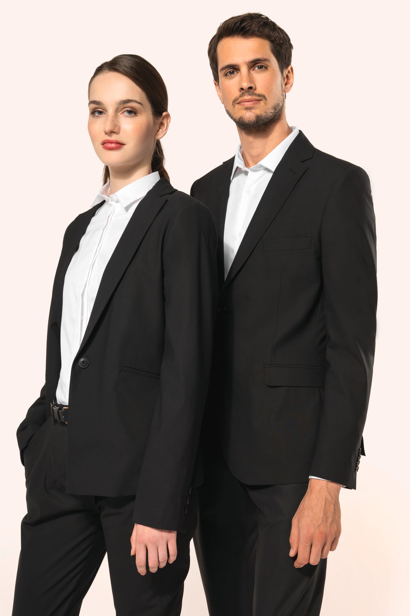 Jeune homme aux cheveux châtains courts et jeune femme aux cheveux châtains longs attachés en queue de cheval composent un duo élégant en noir et blanc. Ils portent chacun une veste noire à la coupe ajustée, associée à une chemise blanche, un pantalon noi