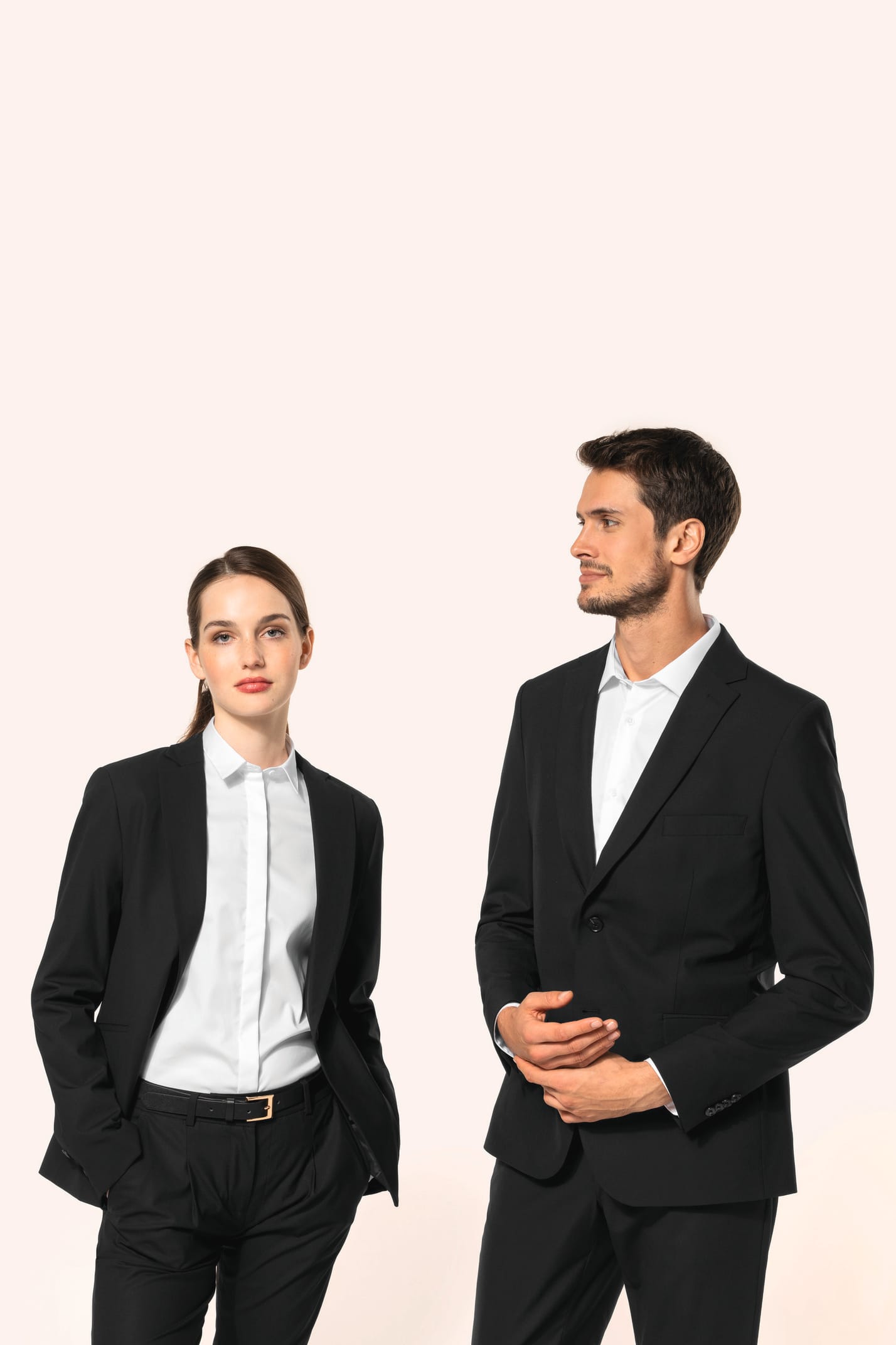 Jeune homme aux cheveux châtains courts et jeune femme aux cheveux châtains longs attachés en queue de cheval composent un duo élégant en noir et blanc. Ils portent chacun une veste noire à la coupe ajustée, associée à une chemise blanche, un pantalon noi