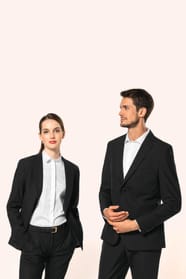 Jeune homme aux cheveux châtains courts et jeune femme aux cheveux châtains longs attachés en queue de cheval composent un duo élégant en noir et blanc. Ils portent chacun une veste noire à la coupe ajustée, associée à une chemise blanche, un pantalon noi