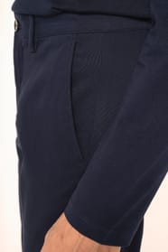 Jeune homme aux cheveux châtains courts portant un chino bleu profond à la coupe straight, moderne et tendance. Confectionné en coton et élasthanne, il associe confort, souplesse et résistance, idéal en toutes saisons. Il l’associe à un T-shirt col V bleu