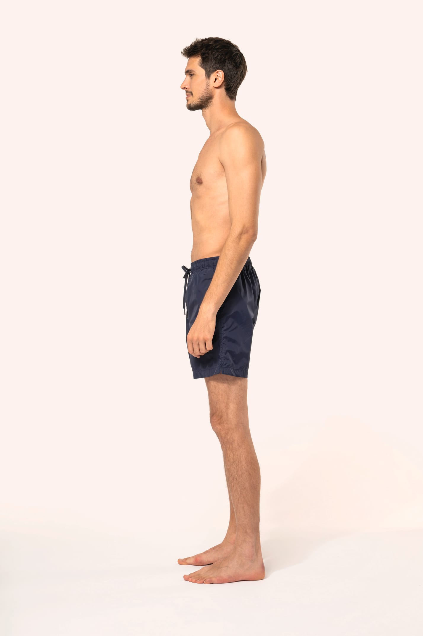 Jeune homme aux cheveux châtains courts et au physique athlétique, torse nu, portant un short de bain en tissu léger. Sa matière favorise un séchage rapide et intègre une poche arrière zippée, combinant confort et praticité pour un style sportif et modern