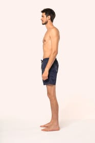 Jeune homme aux cheveux châtains courts et au physique athlétique, torse nu, portant un short de bain en tissu léger. Sa matière favorise un séchage rapide et intègre une poche arrière zippée, combinant confort et praticité pour un style sportif et modern