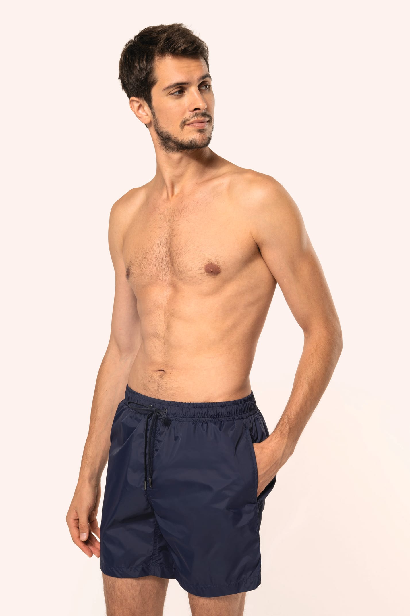 Jeune homme aux cheveux châtains courts et au physique athlétique, torse nu, portant un short de bain en tissu léger. Sa matière favorise un séchage rapide et intègre une poche arrière zippée, combinant confort et praticité pour un style sportif et modern