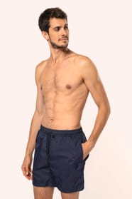 Jeune homme aux cheveux châtains courts et au physique athlétique, torse nu, portant un short de bain en tissu léger. Sa matière favorise un séchage rapide et intègre une poche arrière zippée, combinant confort et praticité pour un style sportif et modern