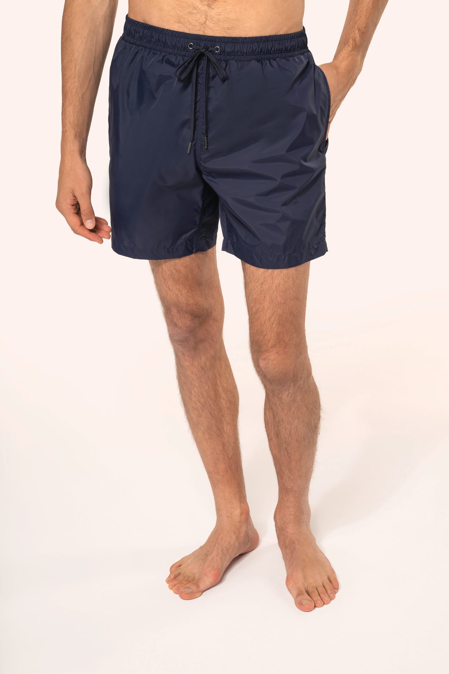 Jeune homme aux cheveux châtains courts et au physique athlétique, torse nu, portant un short de bain en tissu léger. Sa matière favorise un séchage rapide et intègre une poche arrière zippée, combinant confort et praticité pour un style sportif et modern
