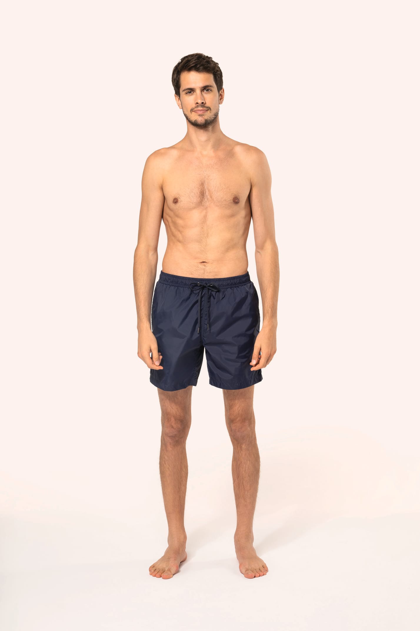 Jeune homme aux cheveux châtains courts et au physique athlétique, torse nu, portant un short de bain en tissu léger. Sa matière favorise un séchage rapide et intègre une poche arrière zippée, combinant confort et praticité pour un style sportif et modern