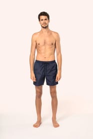 Jeune homme aux cheveux châtains courts et au physique athlétique, torse nu, portant un short de bain en tissu léger. Sa matière favorise un séchage rapide et intègre une poche arrière zippée, combinant confort et praticité pour un style sportif et modern