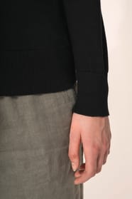 Jeune femme aux cheveux châtains longs attachés en queue de cheval portant un pull noir en coton Supima® col rond. Confectionné en fibres extra-longues, ce coton d’exception offre souplesse, douceur et durabilité. Sa maille révèle des points fantaisie obt