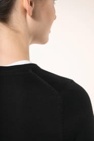 Jeune femme aux cheveux châtains longs attachés en queue de cheval portant un pull noir en coton Supima® col rond. Confectionné en fibres extra-longues, ce coton d’exception offre souplesse, douceur et durabilité. Sa maille révèle des points fantaisie obt
