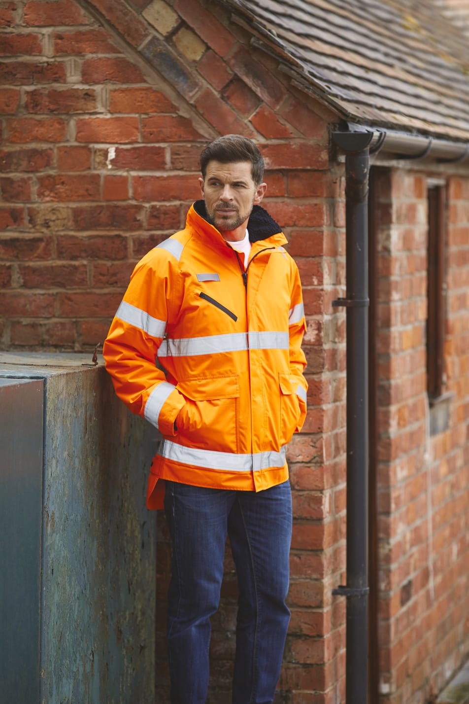 veste orange fluo haute visibilité avec bandes réfléchissantes fullzip multipoches col montant portée par un homme avec les mains dans les poches