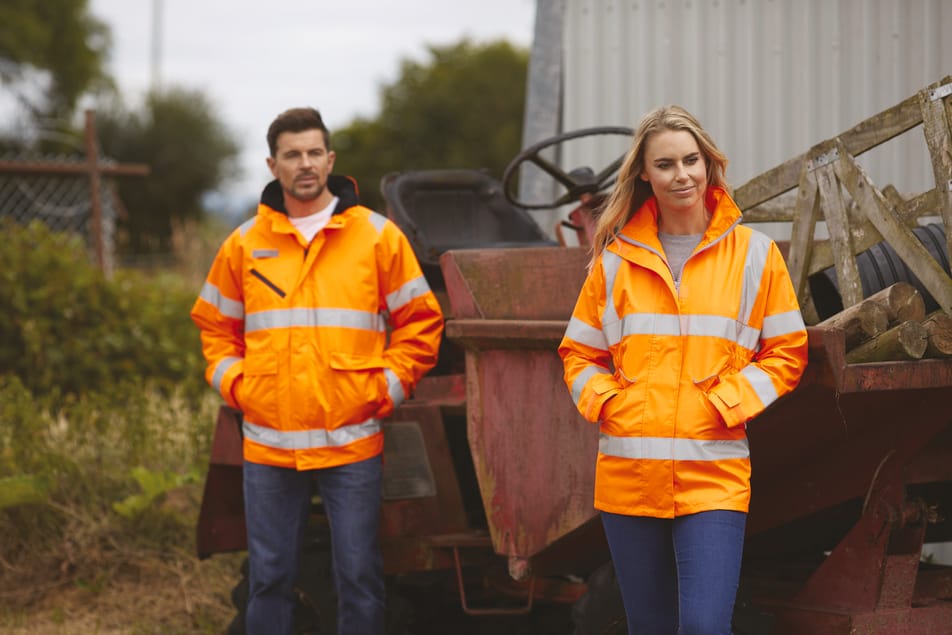 veste orange fluo haute visibilité avec bandes réfléchissantes fullzip multipoches col montant portée par un homme et une femme avec les mains dans les poches