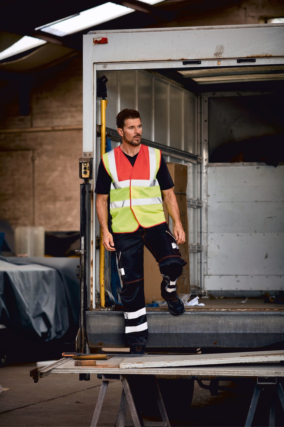 gilet bicolore rouge et jaune fluo haute visibilité avec bandes réfléchissantes sans manches fermeture velcro porté par un homme à l'arrière d'un camion